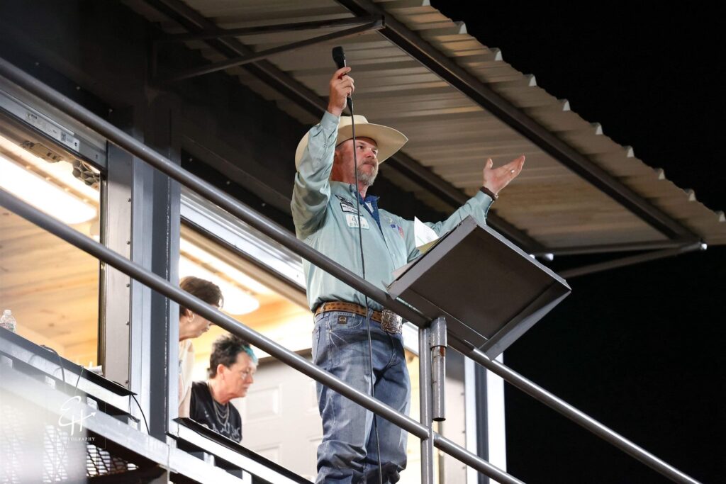 JR McLelland on the announcer&rsquo;s stand at night, raising a microphone with his other arm outstretched toward the arena.