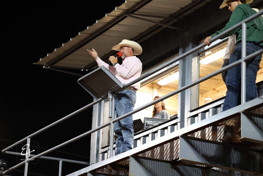 JR McLelland on the announcer&rsquo;s stand at night in a pink shirt, gesturing toward the arena while speaking into a red microphone.