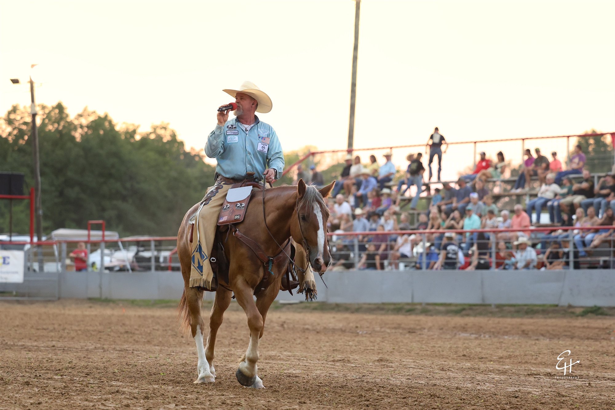JR McLelland riding a sorrel horse with a white blaze, speaking into a red microphone in a rodeo arena, with spectators in the grandstand behind him.