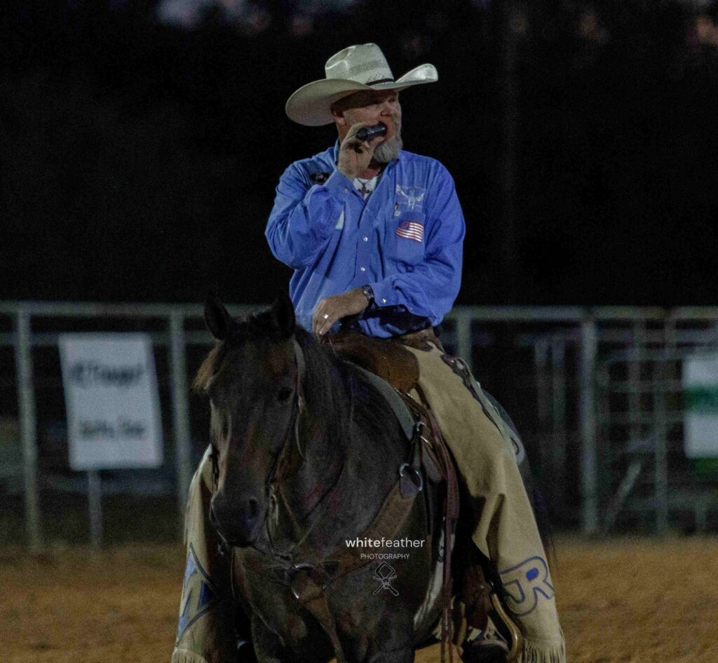 JR McLelland announcing on horseback at night, wearing a blue shirt and chaps.