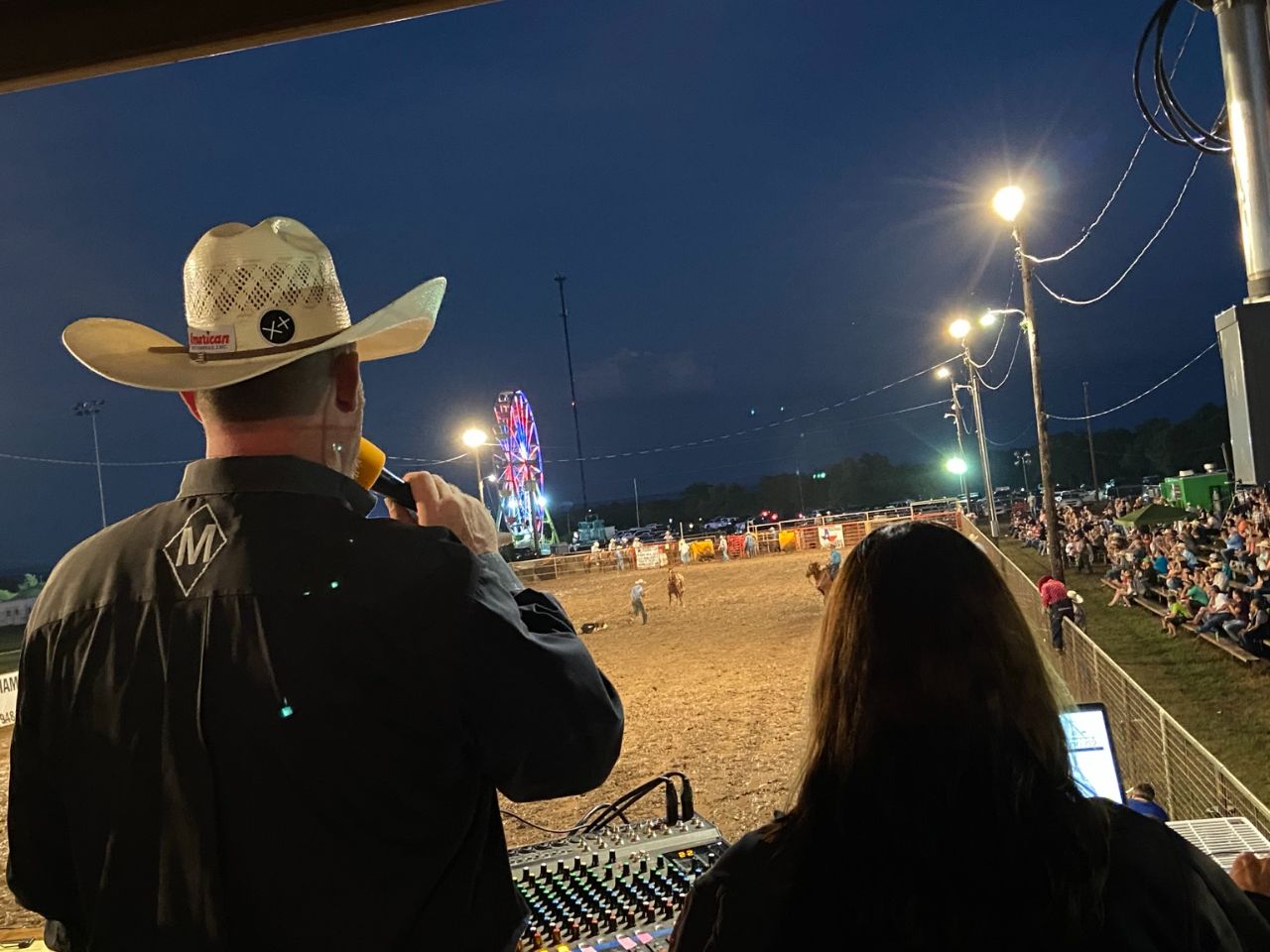 JR McLelland speaking into a microphone beside Michelle at the soundboard, overlooking a rodeo arena at night.